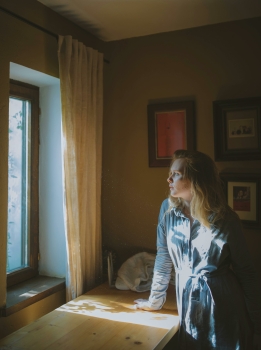 Vrouw staat bij een raam met jas aan en kijkt naar beneden in zacht ochtendlicht, passend bij stress en vermoeidheid.