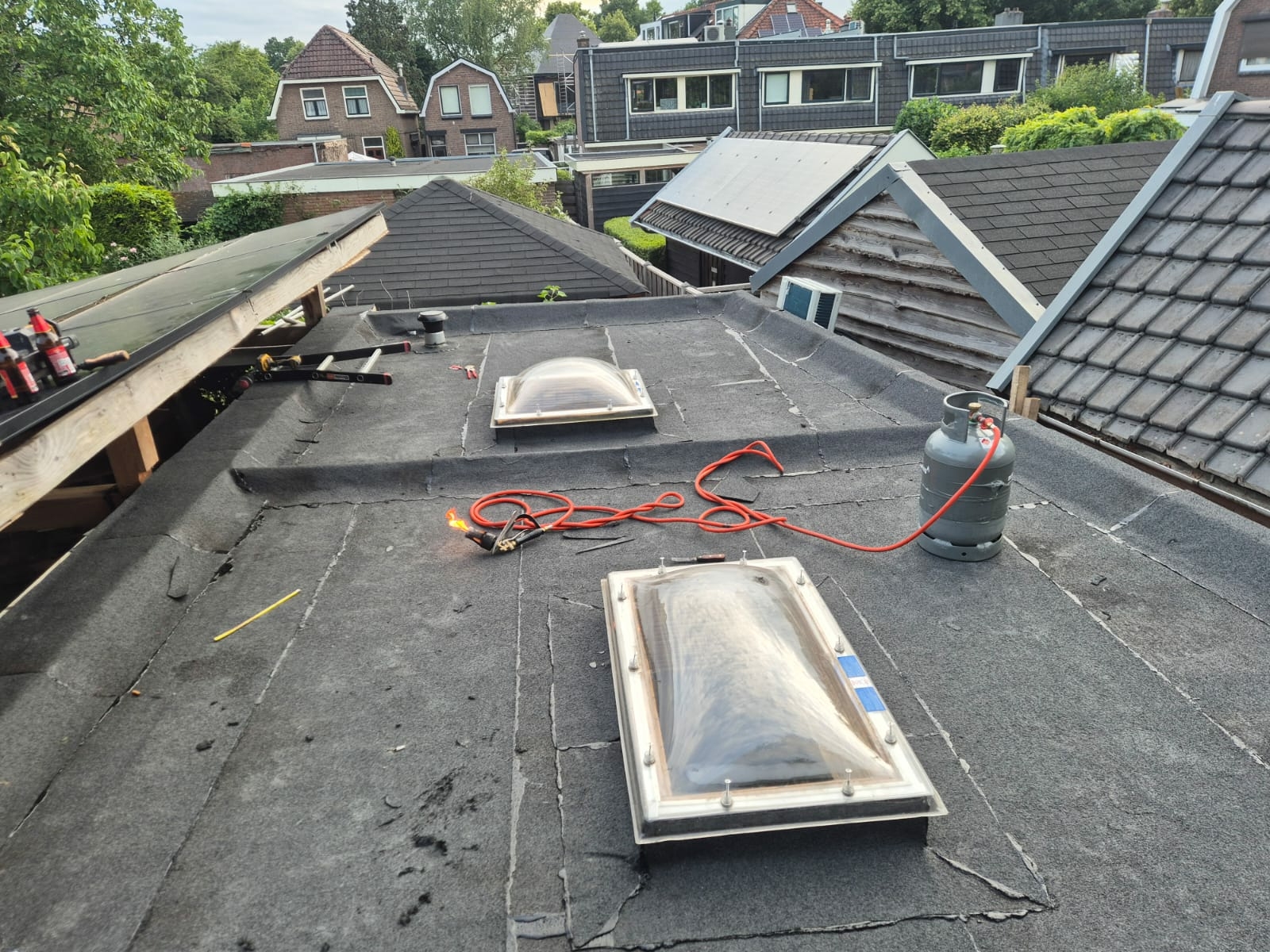 Plat dak met bitumen dakbedekking, twee lichtkoepels en een aangesloten gasbrander — tekenen van slijtage en voorbereidingen voor dakreparatie zichtbaar.
