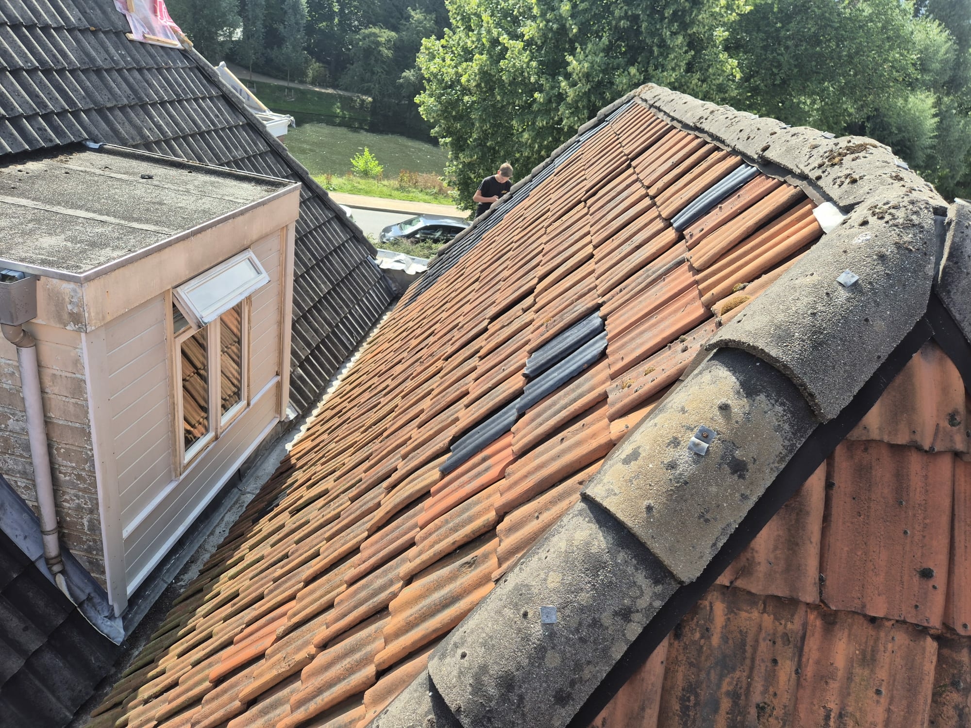 Hellend dak met oranje dakpannen waarvan meerdere zijn vervangen door zwarte noodpannen, teken van recente dakreparatie of lekkageproblemen.