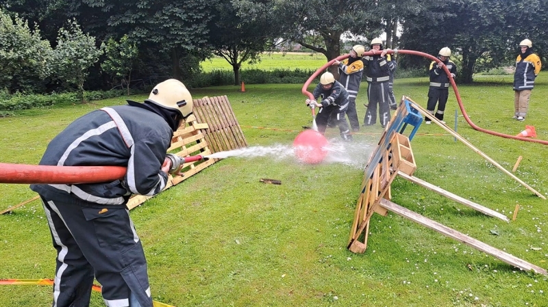 Team collega's in brandweerpakken spuit met een brandslang op een rode bal tijdens een blus-challenge; uniek brandweer-bedrijfsuitje nabij Oss (Heerewaarden), ideaal voor teambuilding en teamwork.