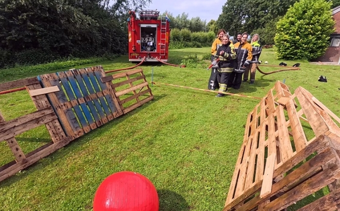 Deelnemers spuiten met een brandslang om een grote rode bal door een palletparcours te duwen; brandweerauto op de achtergrond tijdens het familiebijeenkomst arrangement.