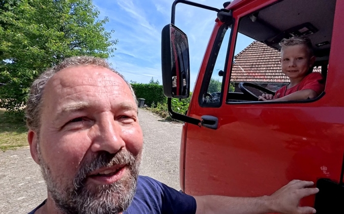 Volwassene staat naast de rode brandweerauto terwijl een kind lachend achter het stuur zit tijdens het familiedag arrangement.