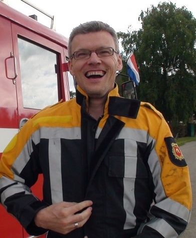 Lachende brandweer-instructeur in pak voor een rode brandweerauto met Nederlandse vlag op de achtergrond; geschikt beeld voor bedrijfsuitje en korte teambuilding met brandweer in Heerewaarden (ook voor Den Bosch, Zaltbommel en Tiel).