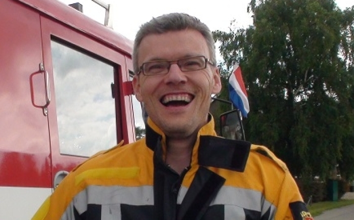Volwassene lacht in brandweerpak naast de rode brandweerauto; activiteit tijdens Opa-en-oma & (klein)kinderdag.