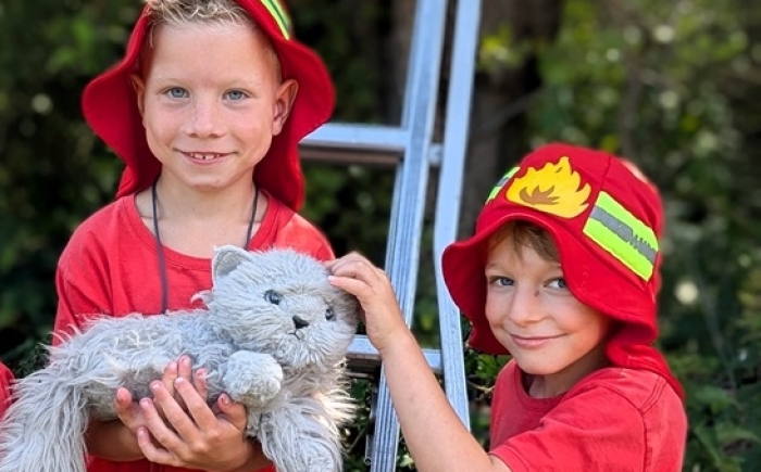 Kinderen in brandweeroutfit poseren bij een ladder met een knuffelpoes; onderdeel van het familiedag arrangement met de brandweerauto.