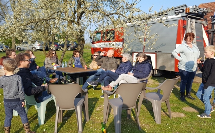 Volwassenen en kinderen zitten buiten onder bloesembomen met drankjes; de brandweerauto staat op de achtergrond tijdens het familiedag arrangement.