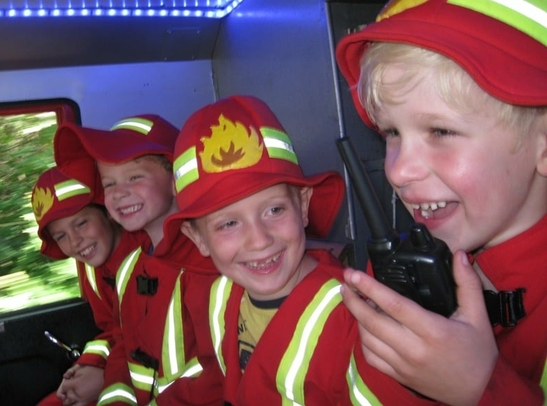 Kinderen genieten van een brandweerkinderfeestje in een echte brandweerauto. Eén kind gebruikt een walkie talkie terwijl de anderen lachen en plezier hebben. Perfect voor 5–7 jaar.