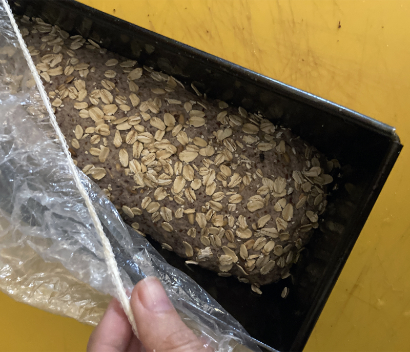 Deeg volkoren meergranenbrood - Marije Bakt Brood in bakblik met hand gele snijplant havervlokken