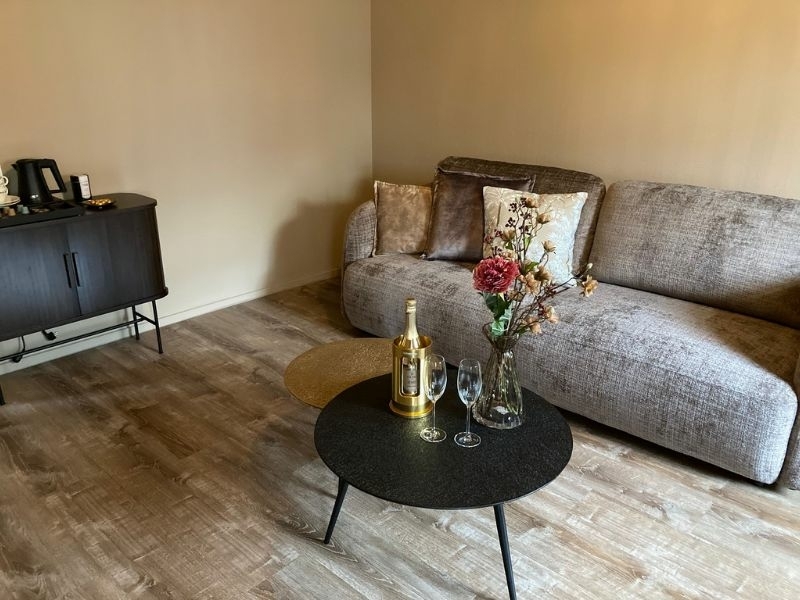 Ruime zithoek met bank, champagne en bloemen in de suite van De La Frontière Bed & Borrelbistro in Slenaken