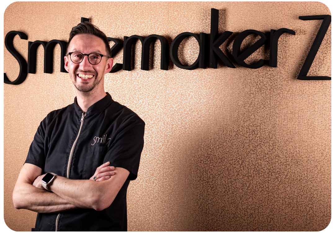 Portret van tandprotheticus bij SmemakerZ praktijk Professioneel portret van een glimlachende tandprotheticus met bril en donkere praktijkkleding, staand met de armen over elkaar voor een wand met het logo van SmemakerZ. De afbeelding heeft een warme en moderne uitstraling.