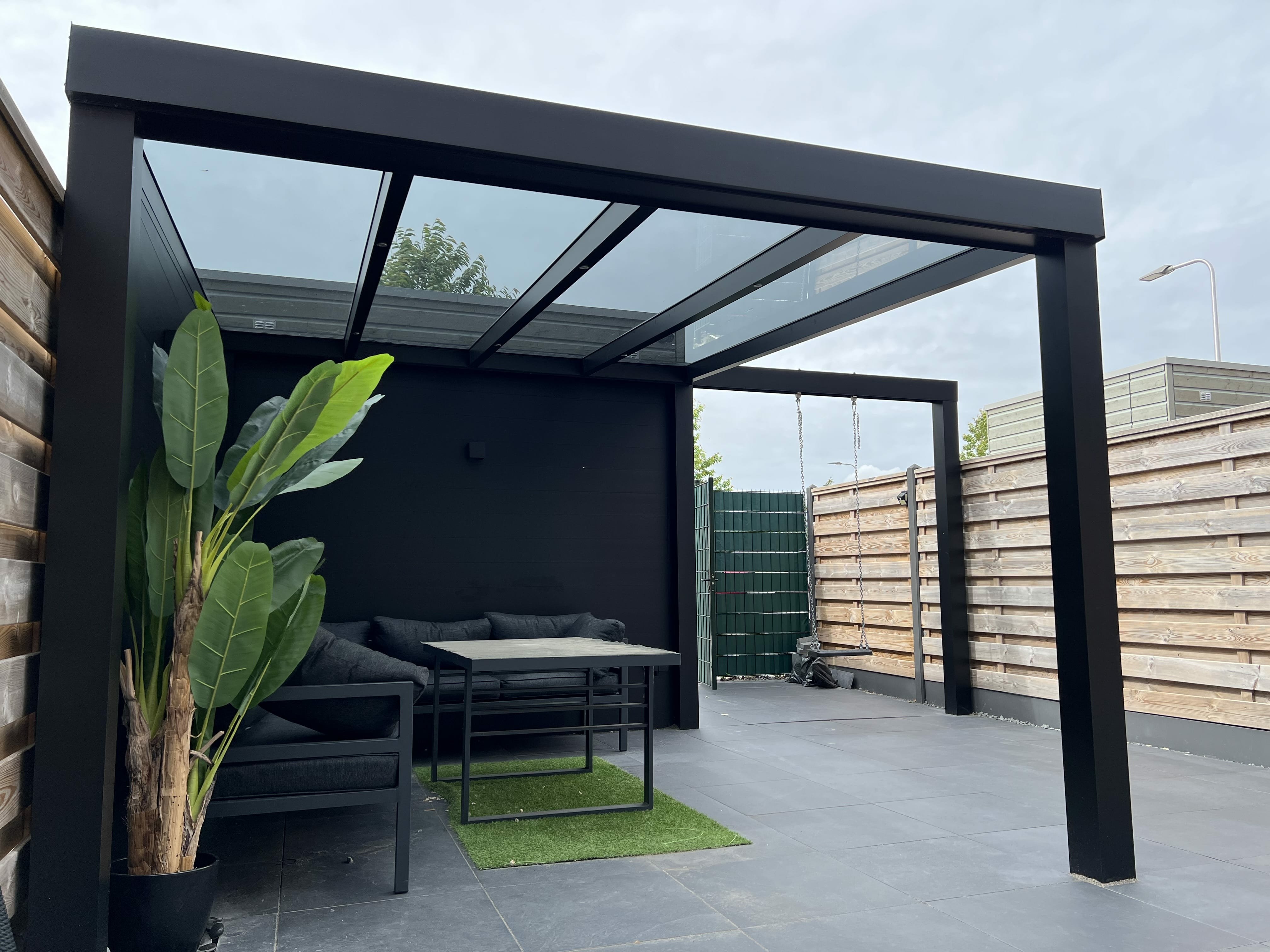 Foto van een moderne aluminium vrijstaande terrasoverkapping met aluminium zijwanden in Lansingerland.. Deze overkapping is uitgevoerd in zwart en voorzien van geïntegreerde ledverlichting. Strakke zwart aluminium overkapping met loungehoek
