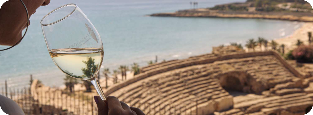 Een vrouw drinkt witte wijn met op de achtergrond het Romeinse Amfitheater van Tarragona aan de kust.