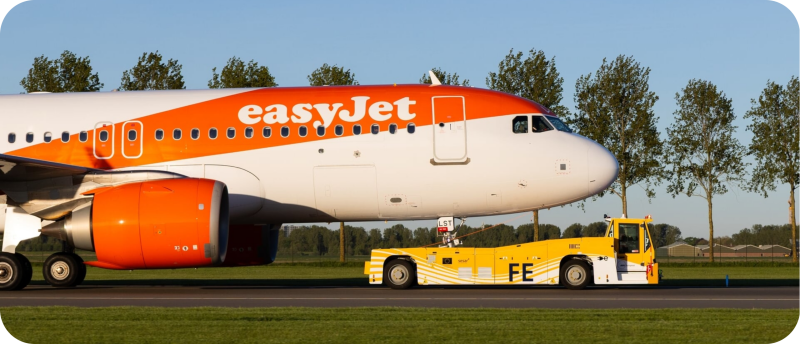 Een oranje-wit easyJet-vliegtuig wordt op Schiphol voortgetrokken door een geel-wit elektrisch TaxiBot-voertuig dat is gekoppeld aan het neuswiel.