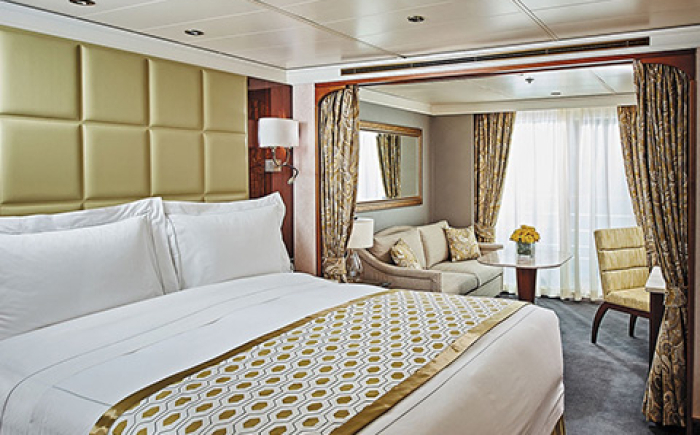 Luxe cruiseschip-suite met groot bed, zithoek, eettafel en balkon. Elegant ingericht met warme tinten en veel natuurlijk licht.