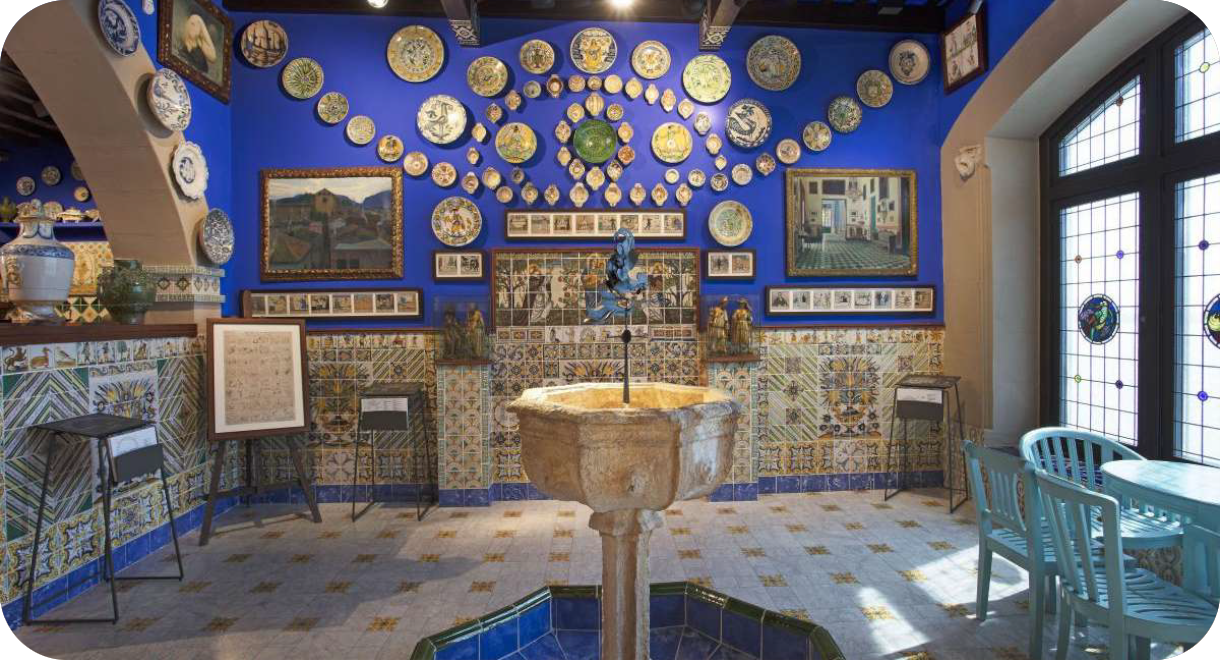 Een interieurfoto van de Sala del Brollador in het Cau Ferrat Museum, met intens blauwe muren, een centrale stenen fontein en wanden versierd met talloze keramieken borden en historische tegels.