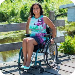 Een jonge vrouw, geïdentificeerd als Claudia, zit in een rolstoel en lacht naar de camera. Ze draagt een roze top en blauwe jeans