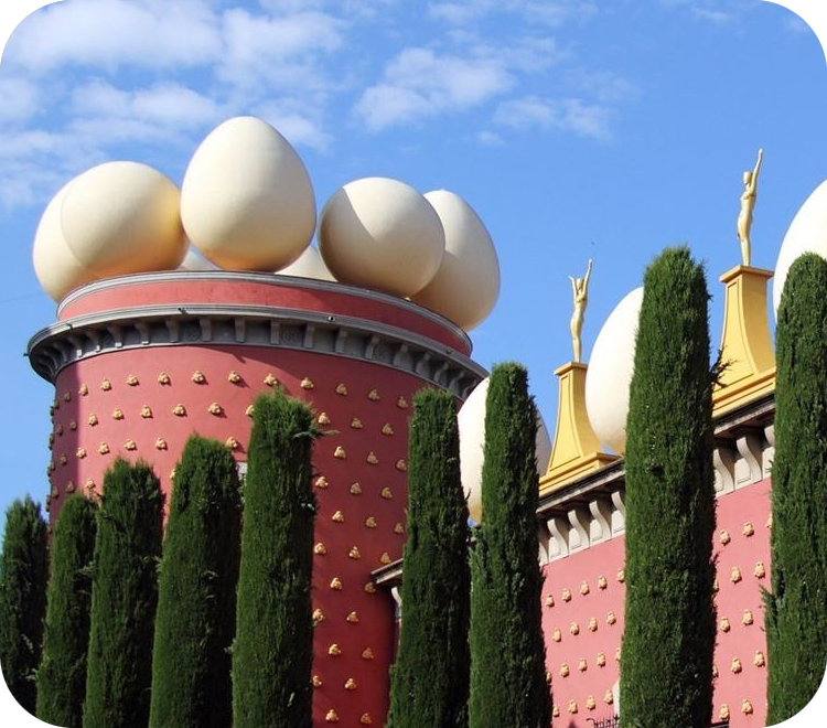 Het rode Teatre Museum Dalí in Figueres met grote witte eieren en gouden beelden op het dak onder een blauwe hemel.