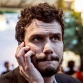 Een close-up van een man met donker, krullend haar en een baardje, die peinzend kijkt terwijl hij zijn hand bij zijn gezicht houdt.