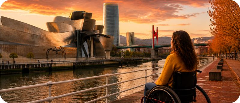Een vrouw in een rolstoel kijkt vanaf een kade uit over de rivier in Bilbao, met op de achtergrond het Guggenheim Museum, de reuzenspin Maman en de Torre Iberdrola tijdens zonsondergang.