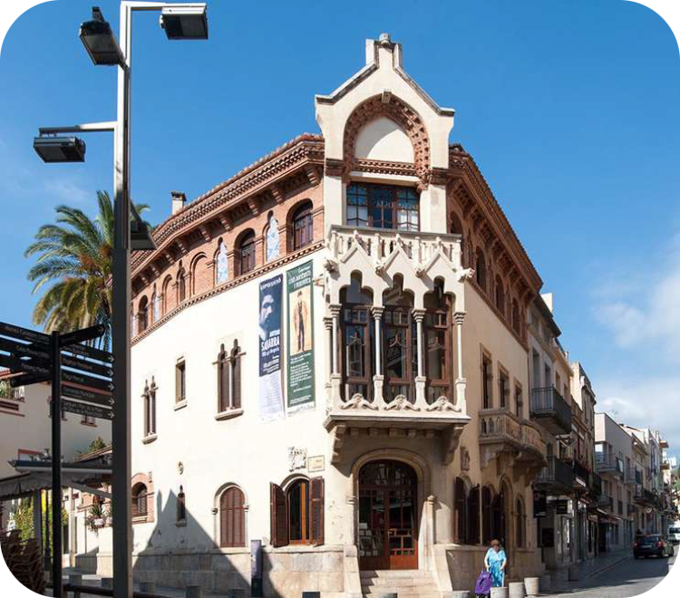 Hoekgevel van het modernistische Casa Museu Lluís Domènech i Montaner in Canet de Mar, Spanje, met een karakteristieke stenen tribune, sierlijke bogen en een blauwe lucht op de achtergrond.