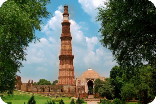 De Qutb Minar, een hoge rood zandstenen minaret met vijf verdiepingen en gedetailleerd snijwerk, omringd door ruïnes en bomen onder een blauwe lucht in Delhi.