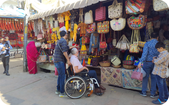 Kleurrijke kraam op een Indiase markt vol handgemaakte, geborduurde tassen en textiel. Tussen de winkelende mensen een man in een rolstoel
