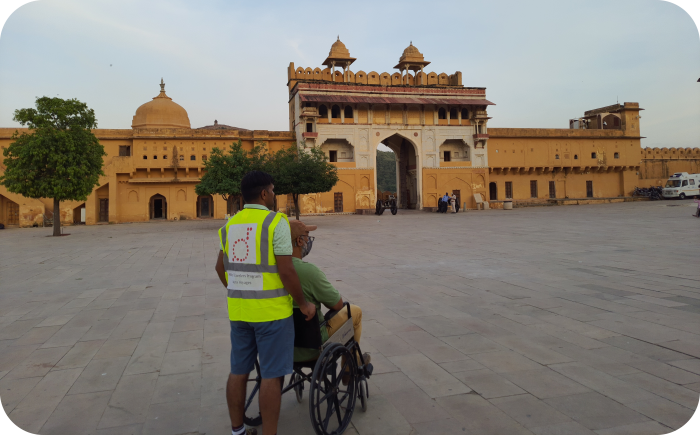 Een man in een rolstoel en zijn begeleider op de zandstenen binnenplaats van het Amer Fort in Jaipur, India, met de monumentale Suraj Pol (Zonnepoort) op de achtergrond onder een heldere lucht.