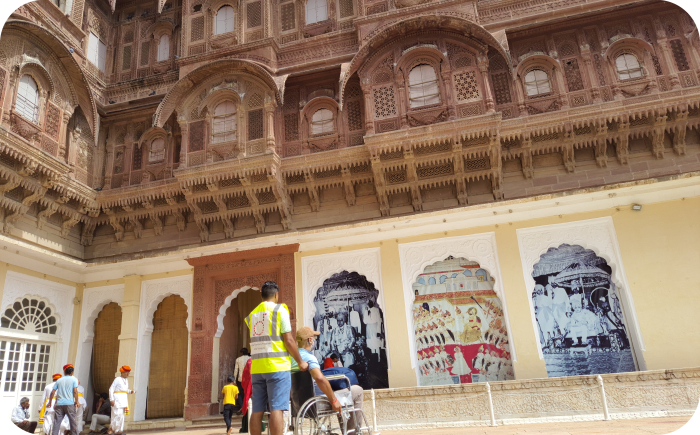 Binnenplaats van het Mehrangarh Fort met een bezoeker in een rolstoel, rijk bewerkte zandstenen balkons en kleurrijke muurschilderingen.