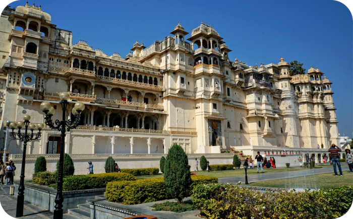 Een groot, wit paleiscomplex (Stadspaleis van Udaipur) met gedetailleerde bogen en koepels onder een blauwe lucht, omgeven door een tuin met groene struiken en lantaarnpalen.