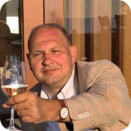 Man in beige jasje zit buiten op een terras in de zon en heft glimlachend een glas rosé richting de camera tijdens een ontspannen reiservaring – *rolstoelvriendelijk reizen*.