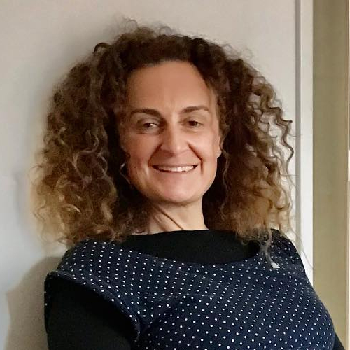 Vrouw met volle krullen, glimlachend, gekleed in een donkerblauwe top met witte stippen, poserend voor een lichtgekleurde achtergrond.