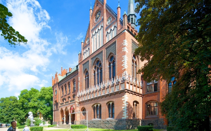 Een neogotisch gebouw van rode baksteen, de Kunstacademie van Letland, staat centraal in het beeld, omringd door bomen onder een blauwe lucht.