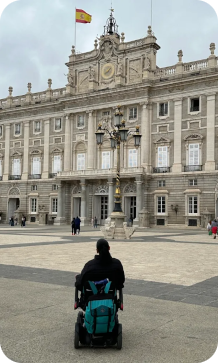 Een persoon in een rolstoel kijkt vanaf een groot plein naar de voorgevel van het indrukwekkende Koninklijk Paleis van Madrid onder een lichtbewolkte hemel.