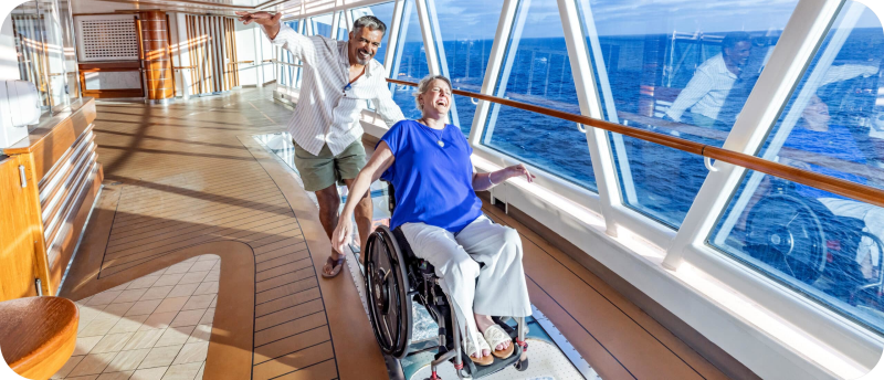 Een vrolijk stel op het dek van een cruiseschip, waarbij een man lachend een vrouw in een rolstoel voortduwt over een glazen vloer met uitzicht op de blauwe oceaan