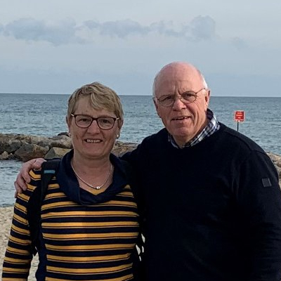 Man en vrouw poseren samen glimlachend aan zee, met rotsen en water op de achtergrond zij draagt een gestreepte trui, hij een donkere trui.