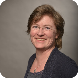 De afbeelding toont een lachende vrouw van middelbare leeftijd met kort, bruin haar en een bril. Ze draagt een donkerblauw of zwart vest over een grijze top en staat voor een effen grijze achtergrond.