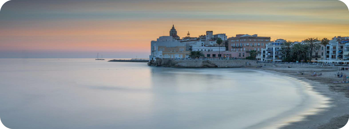 Platja de Sant Sebastia in Sitges bij zonsondergang, met op de achtergrond de historische kerk aan het rustige zeewater.