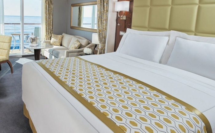 Luxe cruiseschip-suite met groot bed, zithoek, eettafel en balkon. Elegant ingericht met warme tinten en veel natuurlijk licht.