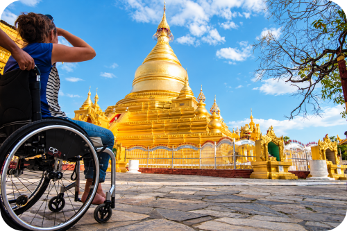 Vrouw in rolstoel bekijkt een indrukwekkende gouden pagode in Myanmar onder een blauwe lucht, terwijl iemand haar rolstoel begeleidt over het stenen plein.