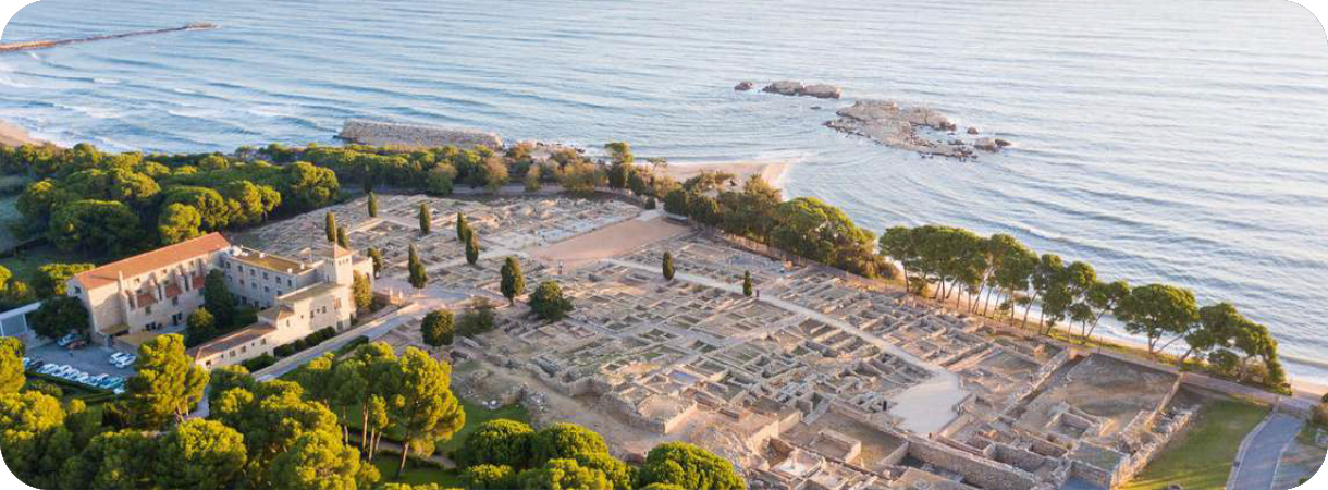 Luchtfoto van de archeologische ruïnes van Empúries aan de kust van de Middellandse Zee in Spanje, omgeven door groene bomen en zandstranden.