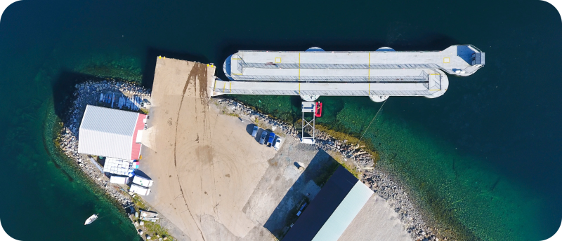 Een luchtfoto van de SeaWalk in Lakselv, Noorwegen: een lange, zilvergrijze inklapbare drijvende cruise-pier die in een S-vorm is opgevouwen aan een landtong in het heldere, groenblauwe water.