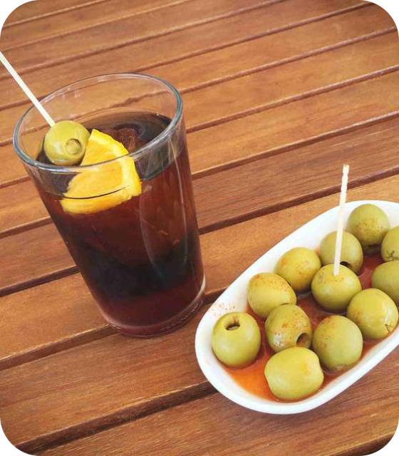 Een glas rode Vermouth met een schijfje sinaasappel en een olijf, geserveerd op een houten tafel naast een schaaltje groene olijven