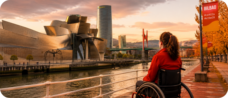Een vrouw in een rolstoel kijkt vanaf een kade uit over de rivier in Bilbao, met op de achtergrond het Guggenheim Museum, de reuzenspin Maman en de Torre Iberdrola tijdens zonsondergang.