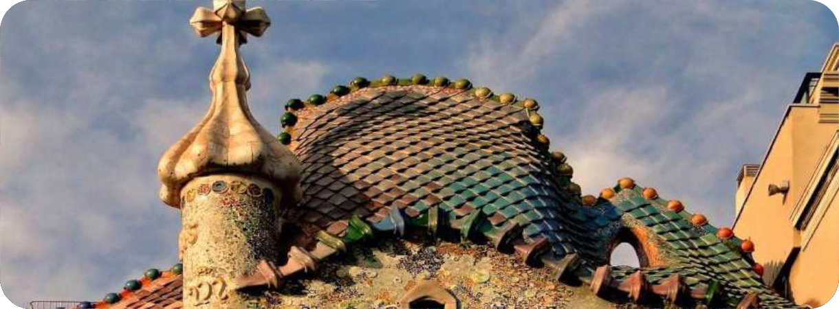 Een close-up van het kleurrijke, golvende dak van Casa Batlló in Barcelona, met glinsterende keramische tegels die lijken op de schubben van een draak en een groot vierarmig kruis op een torenspits tegen een blauwe lucht.