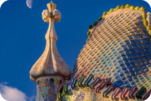 Kleurrijk golvend dak op Casa Batlló met schubben en een witte toren met een kruis tegen een blauwe lucht met een halve maan.
