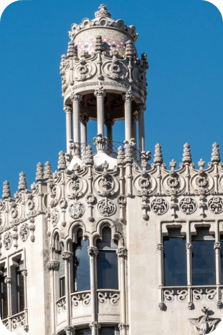 Een gedetailleerde weergave van de rijk versierde, modernistische top en de tempelvormige daktoren van Casa Lleó Morera in Barcelona tegen een helderblauwe lucht.