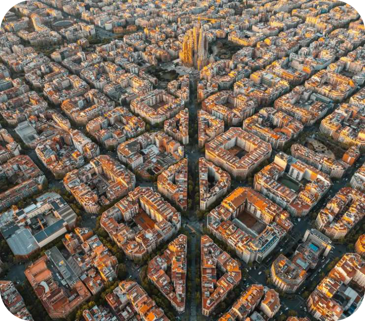Luchtfoto van de wijk L'Eixample in Barcelona met een strak rasterpatroon van vierkante huizenblokken met afgeschuinde hoeken, doorkruist door een diagonale straat, met op de achtergrond de Sagrada Família.