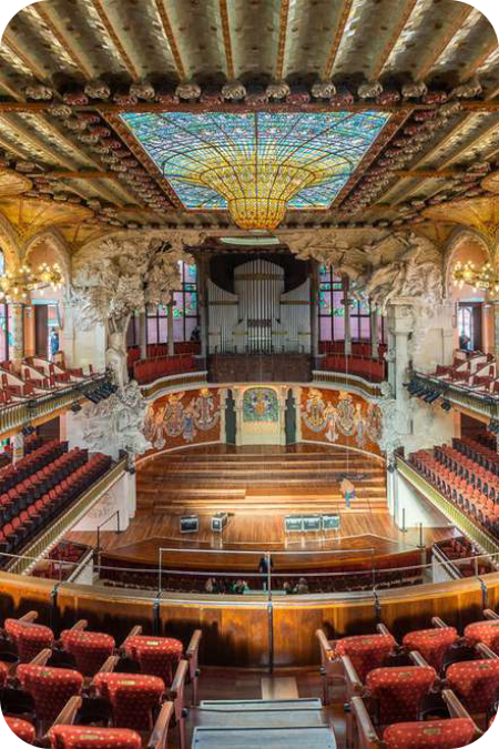 nterieur van de concertzaal van het Palau de la Música Catalana in Barcelona, met een gedecoreerd podium, rijen rode stoelen en een opvallende, kleurrijke glazen lichtkoepel in het plafond.