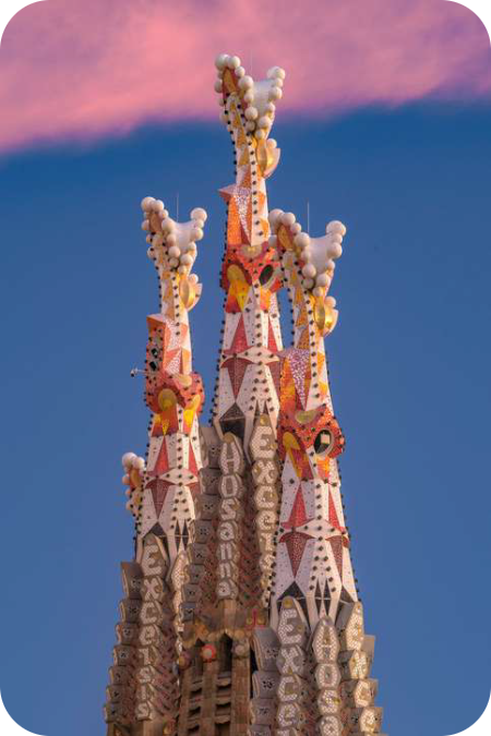 Kleurrijke, met mozaïek versierde pinakels van de Sagrada Família-torens tegen een roze-blauwe avondlucht, met de Latijnse tekst 