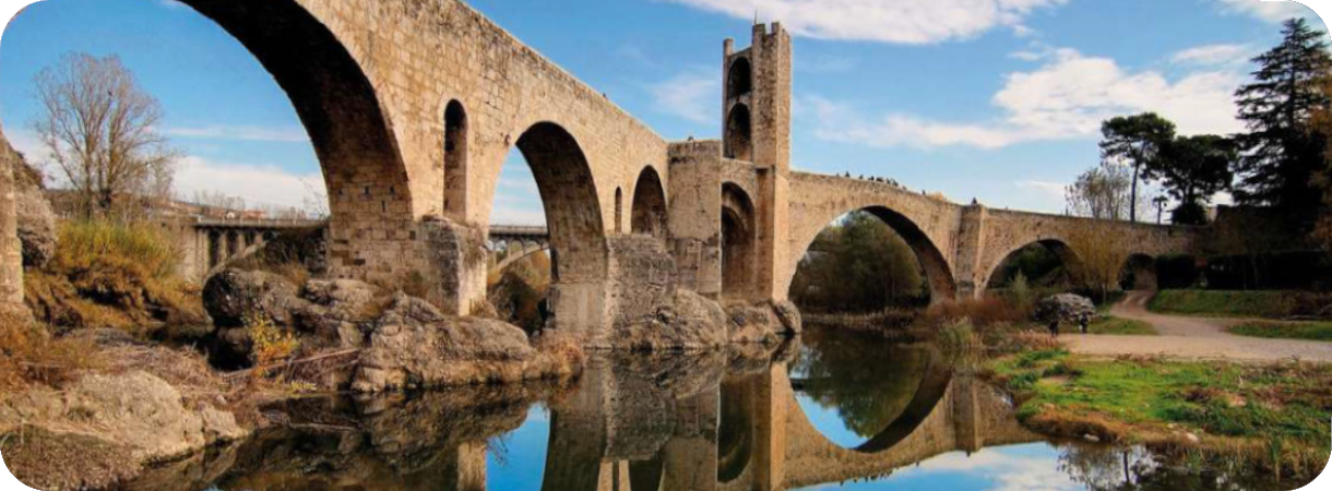 Panoramisch uitzicht op de middeleeuwse stenen brug van Besalú (Spanje) met bogen en een verdedigingstoren, weerspiegeld in de kalme Fluvià rivier onder een blauwe lucht met wolken.